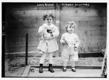 photo en noir et blanc de deux enfants debout habillés en blanc, l'un brun, l'autre, plus petit, blond