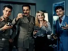 Quatre personnes tenant des verres en main et regardant la caméra. De gauche à droite : deux hommes portant des costumes militaires, une femme aux cheveux blonds et un homme en costume de soirée.