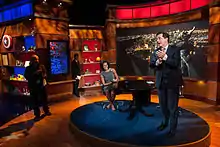 La Première dame Michelle Obama, hilare, assise sur le plateau du Colbert Report et Stephen Colbert est debout sur le plateau, en train de parler et d'applaudir.