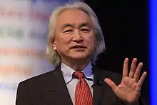 Photo en couleur. Homme asiatique aux cheveux blancs descendants, portant veston et montrant sa main doigts ouverts.
