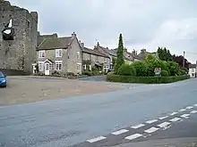 Plusieurs maisons aux murs et aux toits gris derrière un parking et un square. Une route goudronnée traverse l'image au premier plan.