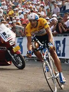 Photographie couleur d'un cycliste portant le maillot jaune et arc-bouté sur son vélo, dans un virage, lors d'un contre-la-montre. Une foule de spectateurs constitue l'arrière-plan
