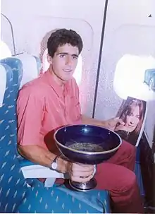 Photographie d'un homme assis dans un avion tenant une coupe dans sa main droite et un magazine dans sa main gauche.