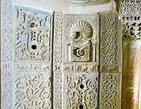 Photographie montrant quelques-uns des panneaux en marbre blanc, sculptés et ajourés, de la paroi du mihrab. Entre les panneaux est visible une partie de l'inscription coranique en caractères coufiques.