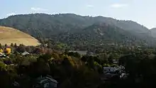 Photo couleur d'une zone boisée, parsemée de quelques bâtiments. Le second plan est composé de collines boisées, sous un ciel bleu nuageux.