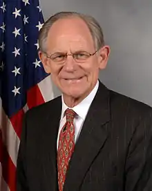 Un homme âgé, portant des lunettes, un costume gris anthracite à lignes, une chemise blanche et une cravate rouge, devant un drapeau américain.