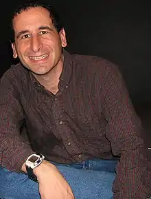 Photographie d'un homme assis. Il sourit, porte une chemise marron et un jean bleu.