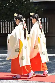Photo couleur de deux jeunes femmes de trois-quart dos, debout, portant un haut blanc, une sorte de jupe longue rouge, et une épingle à cheveux ornée de fleurs au-dessus de leur front.