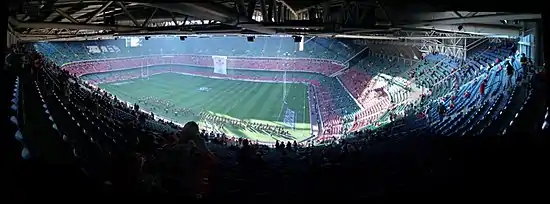 vue panoramique du stade. Juste sous le toit en haut, le photographe montre les tribunes, le stade dans son ensemble. Une fanfare joue alors que peu de public est présent. Les rangées de gradins sont verte ou rouge, par série.
