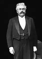 Photo en noir et blanc d'un homme debout, avec les cheveux blancs, une moustache foncée, des lorgnons, un costume trois pièces, un chapeau à la main