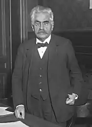 Photo en noir et blanc d'un homme debout dans un bureau : il a des cheveux gris-blancs, d'épais sourcils noirs, une moustache grise, des lorgnons, un nœud papillon et un costume trois pièces