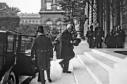 Photo en noir et blanc d'un homme moustachu et à la chevelure blanche sortant d'une voiture et montant les marches d'un bâtiment, avec son chapeau dans la main droite, sous l'objectif d'un photographe et le regard d'autres individus