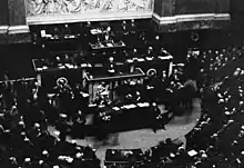 Photo en noir et blanc (vue plongeante) d'un hémicycle, à la tribune de laquelle s’exprime un homme aux cheveux blancs