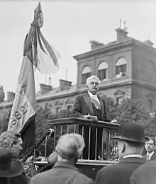 Photo en noir et blanc d'un homme prononçant un discours depuis une élévation en bois située à côté d'un drapeau ; cheveux et moustache blancs, vêtu d'une écharpe élective et d'un costume trois pièces, il tient des feuilles dans sa main droite et s'exprime en regardant son auditoire