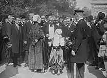 Homme moustachu aux cheveux gris échangeant avec une femme et une fille vêtues de tenues traditionnelles ; en arrière-plan, se trouvent d’autres hommes