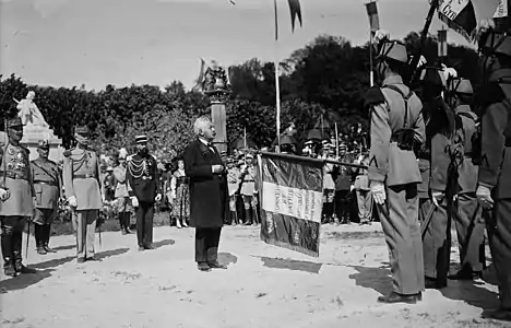 Photo en noir et blanc d'une cérémonie en présence d’un homme en civil, cheveux gris-blancs, moustache grise, nœud papillon, veston et manteau noirs, le regard dirigé vers des militaires, dont un tient un drapeau à l’horizontal