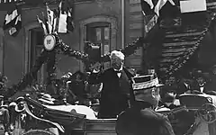 Photo en noir et blanc d'un homme aux cheveux blancs, portant un nœud papillon et un costume trois pièces, en train de saluer la foule avec son chapeau ; en arrière-plan, un bâtiment de la République française est visible