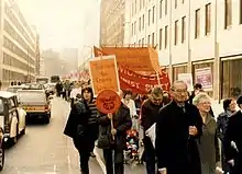 Photographie en couleurs représentant plusieurs personnes manifestent sur la chaussée en brandissant des panneaux en opposition Thatcher.