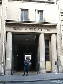 Photo couleur de l'entrée d'un bâtiment, grande ouverte sur un couloir sombre. La façade beiges comprend trois colonnes supportant un fronton sur lequel est inscrit le titre « Ministère de l'Intérieur ». Un homme en uniforme bleu se tient debout devant l'entrée.