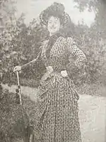 Jeune femme dans un parc avec allée et ombrelle en guise de canne, chapeau, robe longue ancienne.