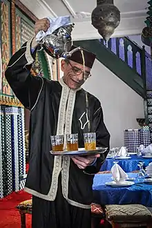 Un homme porte une théière très haut et verse du thé dans des verres posés sur un plateau.