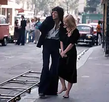 Un homme aux cheveux longs et en costume avec une femme blonde en robe noire et aux pieds nus.