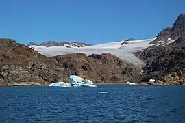 Photographie en couleurs d'un fjord de masses rocheuses brun-foncé, traversées par un glacier et bordées par les eaux d'un fjord.