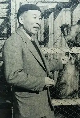 Photo noir et blanc d'un homme âgé, debout de trois quarts. En arrière-plan : deux singes (ç droite) derrière le grillage d'une cage.