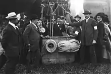 Photographie en noir et blanc d'une femme à bord d'un ballon à air, entourée de nombreux hommes portant des chapeaux.