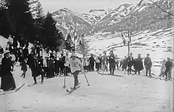 Photographie en noir et blanc d'une concurrente portant le numéro 8 dans une course de ski, devant spectateurs et officiels.