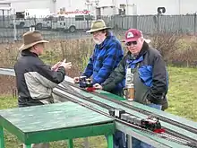 Trois hommes, en tenir pour temps froid, discutent autour d'un ensemble de tables sur lesquelles sont posées trois voies ferrées miniatures. Plusieurs maquettes de trains sont présentes : au premier plan, une locomotive et deux wagons attendent ; à l'arrière plan, une locomotive passe entre les hommes. À l'arrière plan, derrière un grillage, on voit une entrepôt et des automobiles garées.