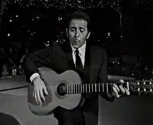 Au centre, sur une scène, un personnage en noir et blanc, buste trois-quart, jouant de la guitare