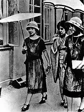 Photo noir et blanc de trois femmes endimanchées, marchant sur un troittoir. Deux, dont une porte une ombrelle, sont habillées d'une robe sombre, la troisième d'un kimono clair et tient un parapluie au-dessus de sa tête.