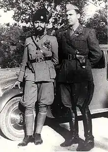 Deux hommes en uniforme - celui de gauche barbu et coiffé d'une toque de fourrure, celui de droite glabre - posant devant une automobile.