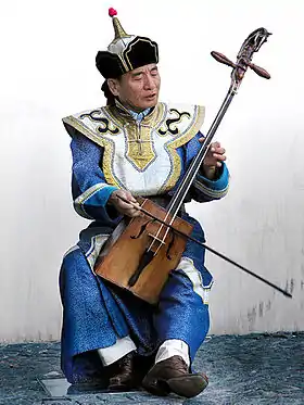 Violoniste en costume traditionnel mongol à dominante bleue, tenant entre ses mains une viole en bois à deux cordes dont le manche se termine en forme de tête de cheval.
