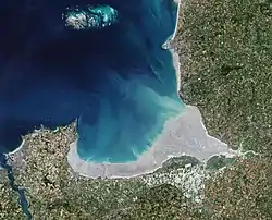 Image satellite de la baie du Mont-Saint-MichelCette image satellite permet d'observer deux paysages différents correspondant à deux grands ensembles morpho-sédimentaires : le domaine occidental, de Cancale à Cherrueix, correspond à un fond de baie abrité. Cet estran large de 5 km, à pente modérée (de 3 à 10 ‰), comprend des vases entaillées par les « biez » (terme local désignant les canaux de drainage du marais de Dol. Le domaine oriental, de Cherrueix à la pointe de Champeaux, correspond à un complexe estuarien au débouché de trois petits fleuves côtiers (la Sée, la Sélune et le Couesnon). Cet estran large de 10 km, très plat, est parcouru par des chenaux de rivière à tendance méandriforme, et un réseau dense de chenaux de marée aux tracés plus rectilignes..