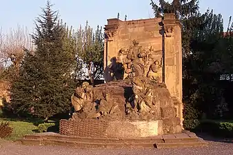 Photographie couleur d'un monument en grès situé en plein air et représentant un Christ debout en prière, entouré de disciples endormis et de soldats en armes.