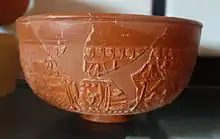 photo couleur d'une poterie rouge restaurée. Les parties reconstituées sont plus sombres et lisses, les morceaux originels comportant frise et personnages.