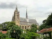Église vue de côté, dépassant des arbres, avec un clocher à quatre tours, et un clocheton au centre du toit d'ardoise.