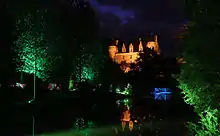 Photographie nocturne en couleurs de jeux de lumières sur le façade d'un château en arrière-plan d'une rivière bordée d'arbres.
