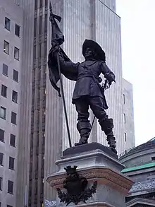 Statue de Maisonneuve portant fièrement un drapeau de la main droite et une épée à la main gauche. Il porte un large chapeau au bord relevé, une tunique, des pantalons et de hautes bottes aux bords repliés.