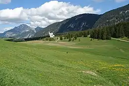 La photo couleur présente un alpage fleuri et vallonné au premier plan. En arrière plan, un monument honorant  la Résistance dans le plateau des Glières se découpe face aux versants boisés et rocheux du fond de l'image.