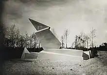 Photographie en noir et blanc d’un monument aux morts. Sculpture en béton aux formes anguleuses.