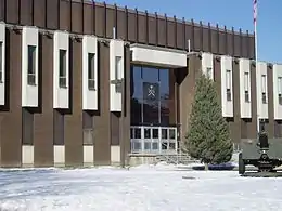 Photo en couleur de la façade d'un bâtiment brun avec une porte vitrée au centre surmontée d'un insigne militaire