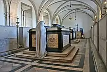 Sépultures du roi et de la reine de Portugal dans une crypte voûtée, les monuments funéraires sont en pierres claires et encadrées d'une armature noire, ils portent deux couronnes sculptées et font face à une statue blanche de pleureuse