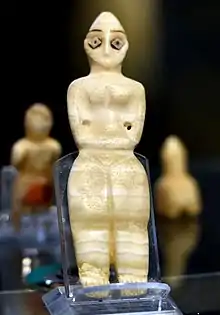 Photo d'une statuette féminine en pierre de couleur beige décorée de morceaux de coquillages collés avec du goudron afin de souligner la présence des yeux.