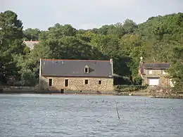 Vue d'un bâtiment en bord d'eau. Le moulin, en pierre, comporte une ouverture basse et quatre ouvertures rectangulaires au-dessus de l’eau.