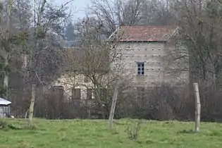 Un moulin à deux étages caché par des arbres sans feuille, une barrière en barbelés est située en prermier plan.
