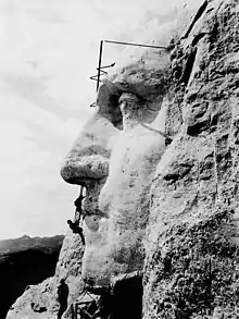 Le visage de George Washington, vu de profil, avec des hommes suspendus à des cordes en train de le sculpter