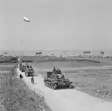 photo montrant une colonne de blindés britanniques avançant dans les terres avec en fond les plages du débarquement